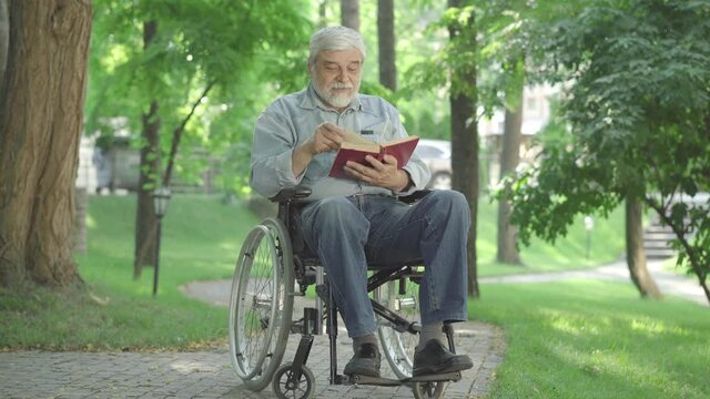 Disabled senior man sitting in wheelchair and reading book. Wide shot portrait of positive Caucasian male invalid enjoying hobby outdoors on sunny summer day. Aging and lifestyle concept.