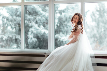 Luxury bride in wedding dress for Christmas near the large panoramic window. Luxury bride in wedding dress for Christmas. Charming bride in an elegant wedding dress. Bride in luxurious suites.