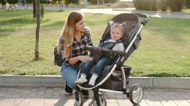 Mother walks with her daughter who is sitting in a stroller in public park. Love and care.