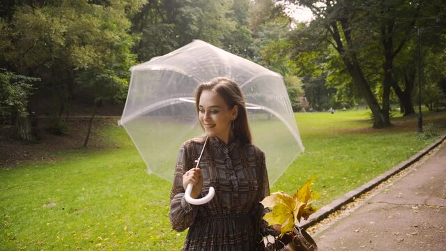 Happy young woman walking outdoors under transparent umbrella during rain. Fall season activities. Stylish girl enjoys autumn weather dancing and having fun.