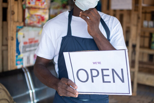 African Business Owner Wearing A Face Mask And Holding An Open Sign In Front Of His Shop