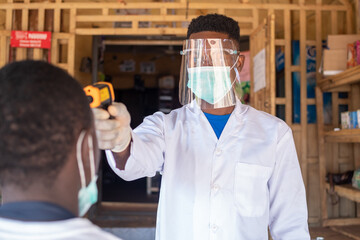 an african man in full protective medical gear from coronavirus using an infrared thermometer in...