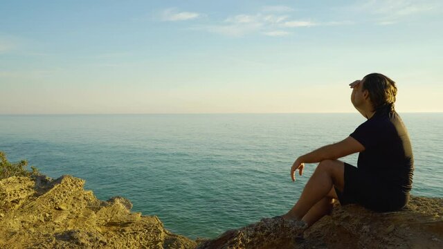 Hombre sentado en un acantilado al atardecer con el mar, el horizonte y el cielo de fondo, mirando, se&ntilde;alando con el dedo y saludando. Desde Conil, C&aacute;diz, Andaluc&iacute;a, Espa&ntilde;a, Europa.