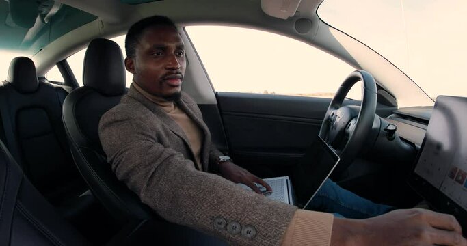 Portrait of happy young African American male traveling in self-driving electric car with autopilot and typing on laptop. Handsome businessman sitting in driverless vehicle and tapping on computer