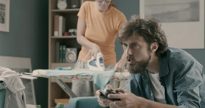 Lazy husband playing videogames while his wife is ironing