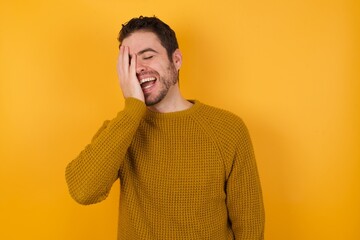 Young man wearing casual sweater and over isolated yellow background makes face palm and smiles...