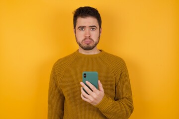 Upset dissatisfied Young man wearing casual sweater and over isolated yellow background uses mobile software application and surrfs information in internet, holds modern mobile hand
