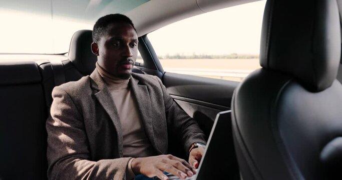 Portrait of happy young African American male traveling in self-driving eco electric vehicle and typing on laptop. Handsome businessman sitting in driverless car with autopilot and tapping on computer