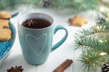 Obraz na płótnie Canvas Mint cup of tea with heart, gingerbread next to branches of a Christmas tree with ight bulbs on white wooden background
