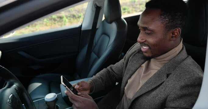 Close up of handsome African American man in electric automobile smiling and searching internet on cellphone. Young happy male sitting in modern car and tapping on smartphone. Business concept