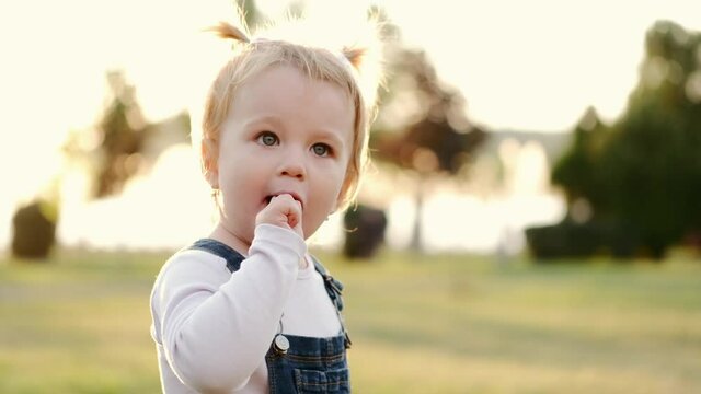 Little girl sucks chupa chups outside in the park. Close-up funny baby with two tails. A girl holding a candy in her hands, takes it in her mouth and licks