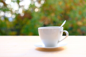 close-up of female hand placing hot ceramic white coffee cup with smoke on saucer over wooden table in nature green background, outdoor morning tea concept, seasonal mood