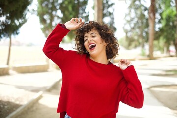 Photo of upbeat Young arab woman wearing casual red sweater in the street has fun and dances carefree wear being in perfect mood makes movements. Spends free time on disco party