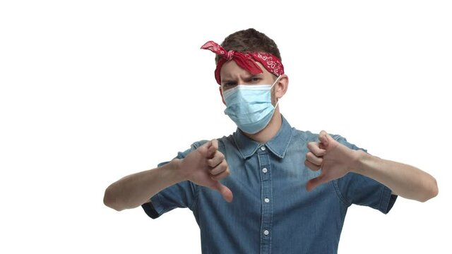 Coronavirus, social distancing and lifestyle concept. Young male hipster with red bandana on forehead, wearing medical mask and shirt, showing thumbs-down, disapprove something bad