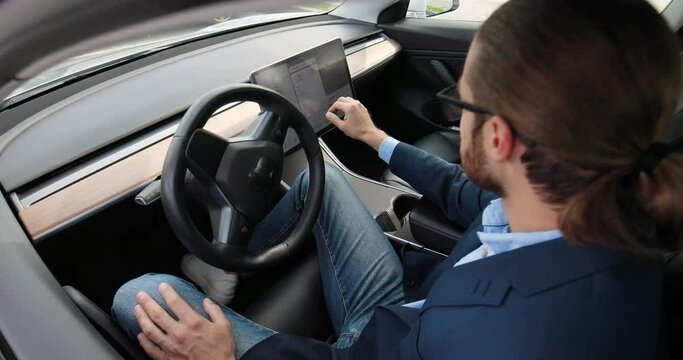 Caucasian man sitting in electric vehicle and typing on on-board touch screen using GPS navigation map. Handsome male driver traveling in futuristic car and tapping on dashboard computer