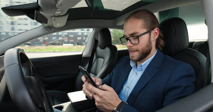 Close up portrait of Caucasian man driver sitting in electric car and smiling while browsing on mobile phone. Young handsome cheerful male in modern fancy car typing on smartphone. Urban concept