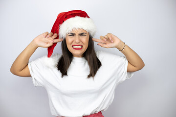 Young beautiful woman wearing a christmas hat over white background covering ears with fingers with annoyed expression for the noise of loud music. Deaf concept.