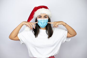 Young beautiful woman wearing a christmas hat over white background pointing the mask. Warning expression with negative and serious gesture on the face.