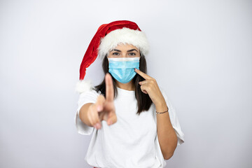 Fototapeta na wymiar Young beautiful woman wearing a christmas hat over white background pointing the mask. Warning expression with negative and serious gesture on the face.