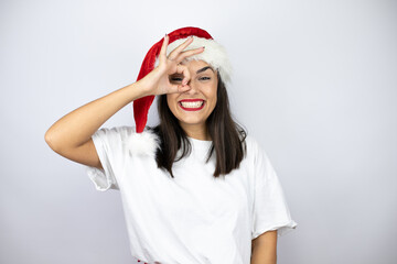 Young beautiful woman wearing a christmas hat over white background doing ok gesture shocked with smiling face, eye looking through fingers