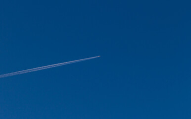 A single plane flies across the clear blue sky leaving a contrail. White jet passenger airplane flying at high altitude in the cloudless sky. Behind the aircraft is white vapor trails.
