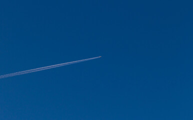 A single plane flies across the clear blue sky leaving a contrail. White jet passenger airplane flying at high altitude in the cloudless sky. Behind the aircraft is white vapor trails.