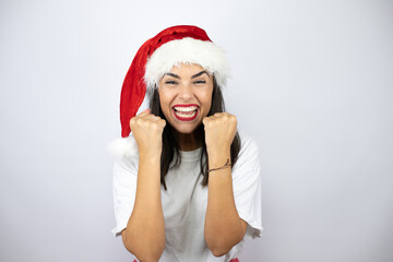 Young beautiful woman wearing a christmas hat over white background very happy and excited making winner gesture with raised arms, smiling and screaming for success.