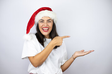 Young beautiful woman wearing a christmas hat over white background surprised, showing and pointing something that is on her hand