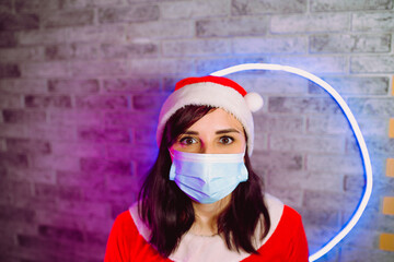 Young woman in medical mask and Santa Claus suit against illuminated wall. Close up of brunette in Christmas hat and protective mask. Concept of safe Christmas celebration during coronavirus pandemic