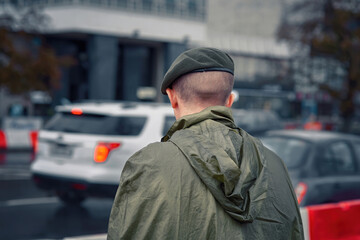 Soldier in green beret and raincoat keep watch in front of a cordon line. Crime prevention and the search for criminals. Military man stand at post. View from the back