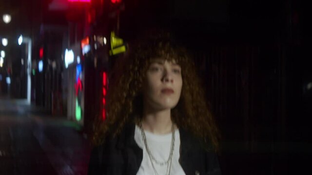 Curly haired woman in white t-shirt and black jacket walks street at the night in anamorphic flares and city lights - Fashion clothing and feminism concept