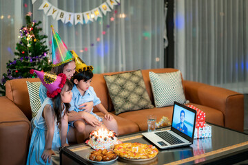 Happy girl and son celebrating birthday with her mother at home with father on video call in laptop while blowing candles in cake on table