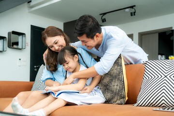 Asian family with father, mother and her daughter reading fairy tail story book on sofa at home together and pointing on the book looking it with happiness spending time together.