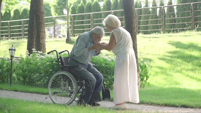 Caring senior woman helping handicapped man to stand up from wheelchair and walk. Thankful Caucasian husband hugging loving wife and smiling. Rehabilitation and support.
