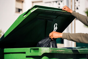 Man throwing out black eco-friendly recyclable trash bag in to big plastic green garbage container....