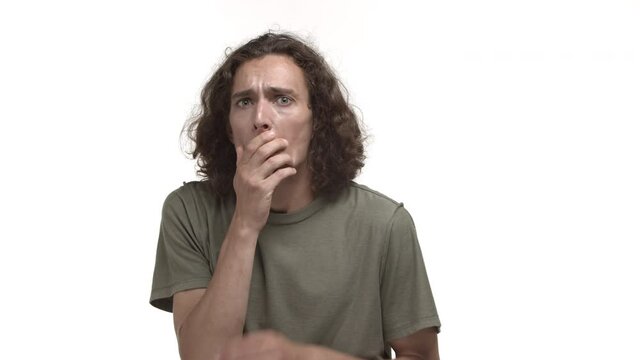 Video of attractive young man with wavy hair, wearing casual t-shirt, grimacing and gasping shocked, reacting to something bad and terrible, standing alarmed over white background