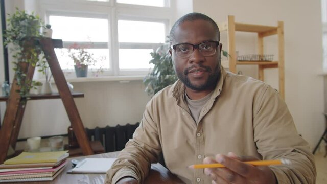 African American male teacher sitting at home, showing educational poster and speaking at camera while giving online lesson via video call during covid lockdown