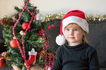 Christmas and New Year holidays preparation concept. Happy smiling child decorating  and looking at Christmas tree, wearing dress and santa hat. 
