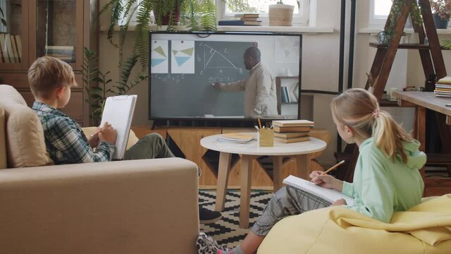 Little brother and sister sitting together in living room, listening to black male teacher via video call on TV and writing in workbooks while studying remotely at home during covid lockdown
