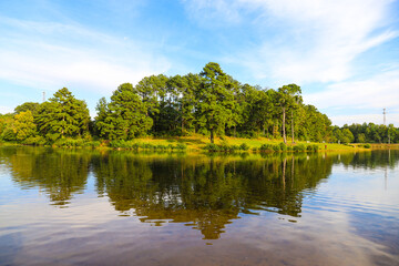 Fototapeta na wymiar stunning shot of the still lakes waters with lush green and autumn colored trees and blue sky and clouds reflecting off the lake at Rhodes Jordan Park at Lawrenceville Lake in Lawrenceville, Georgia