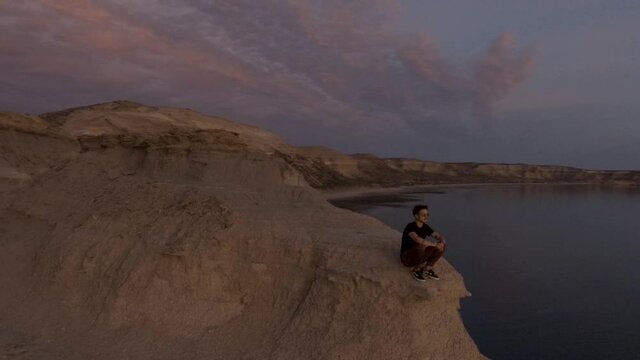 Lonely Young Man Sitting At The Mountain Edge Looking Into The Distance At Sea During Sunset - Aerial Orbital Shot