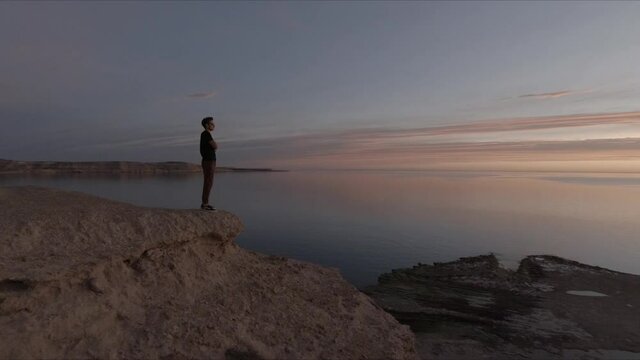Man standing on top of a cliff next to a small village watching the sea at sunset - Aerial orbital shot