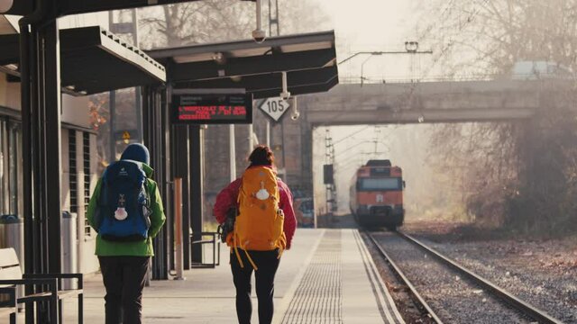 Two People wearing warm jackets beanies and backpacks walk up a European train station as the train departs after getting off