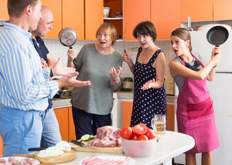 Three women holding frying pans opposing two men in kitchen