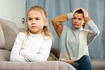 Frustrated small girl sitting at sofa, having conflict with mother at home interior