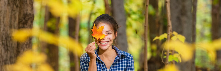 Happy Asian woman holding red maple leaf to her eye cute portrait outdoor nature banner. Panoramic autumn walk in forest.