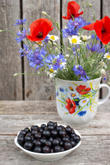 Black currants in small dish and beautiful wild flowers inside tea cup on wooden background