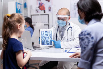 Doctor wearing visor against coronavirus pointing at radiography during consultation of mother and child. Pediatrician specialist with protection mask providing health care service radiographic