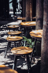 Fototapeta na wymiar Vertical atmospheric warm summer cover photo, wooden stools chairs in outdoor cafe on veranda in garden, bright rays sun beam through trees. Positive good calm mood concept, for wallpaper screensaver