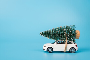 Merry Christamas concept. Side profile close up photo of mini toy white stylish modern car carrying fir tree in snow on the top of the roof isolated over blue color background